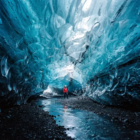 A tiny explorer in a red jacket stands inside a vast glowing blue glacier cave beside a meltwater stream, wide photograph.